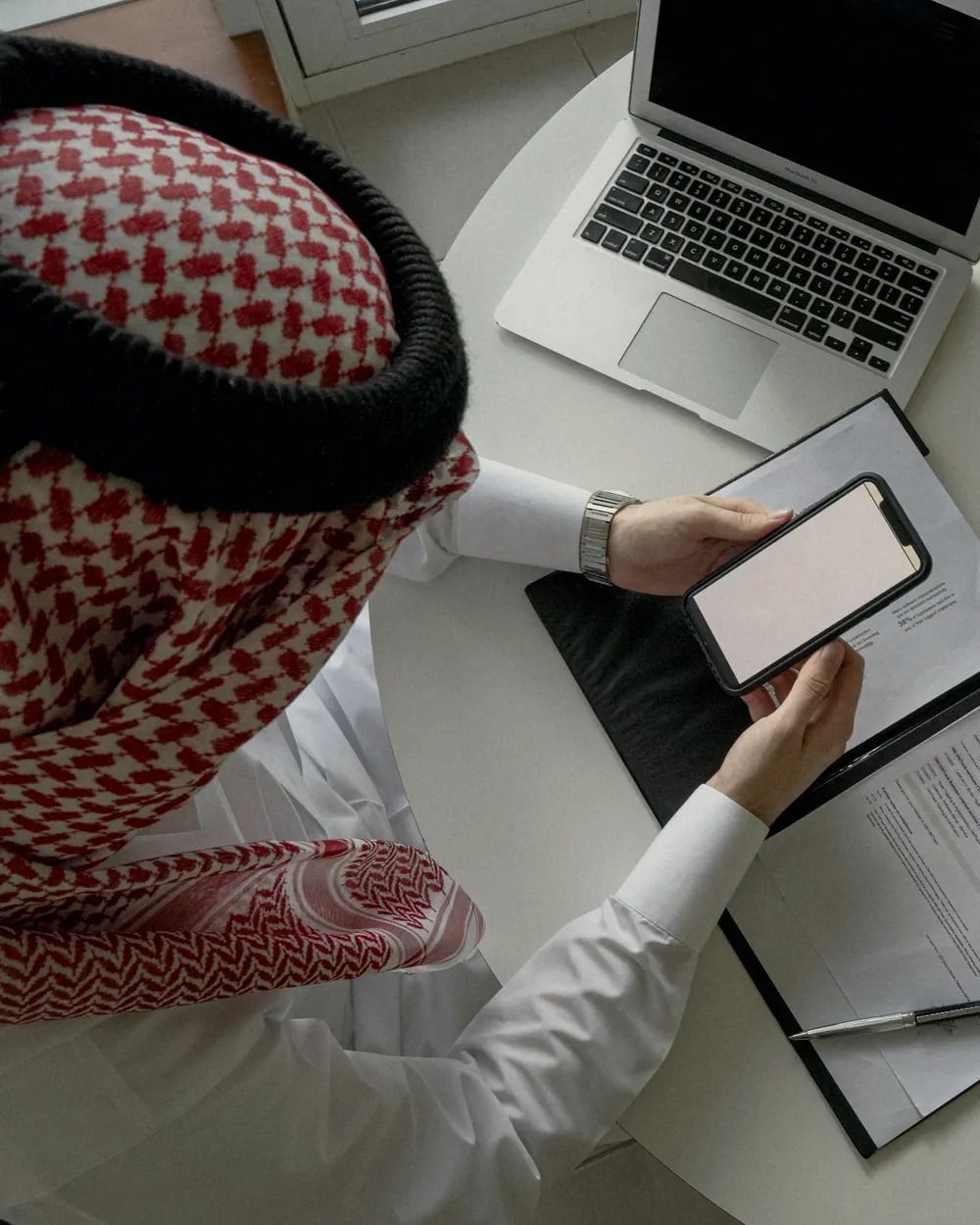 Professional in a shemagh using a smartphone beside a laptop